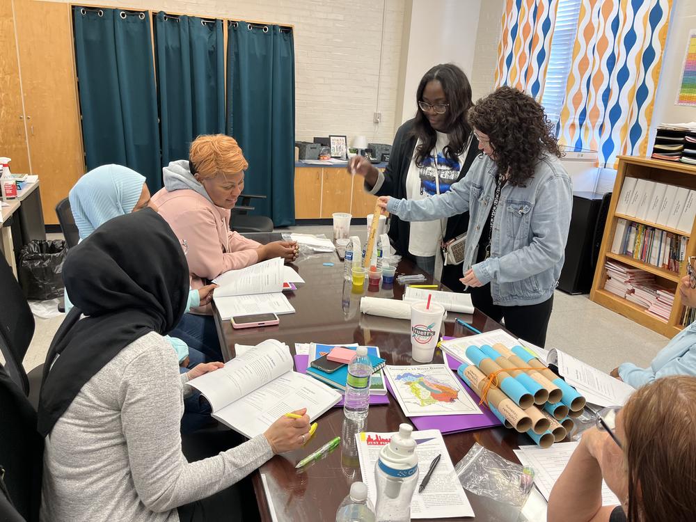 People are gathered around a table with books, cups and other materials scattered about as a demonstration with water is being led.
